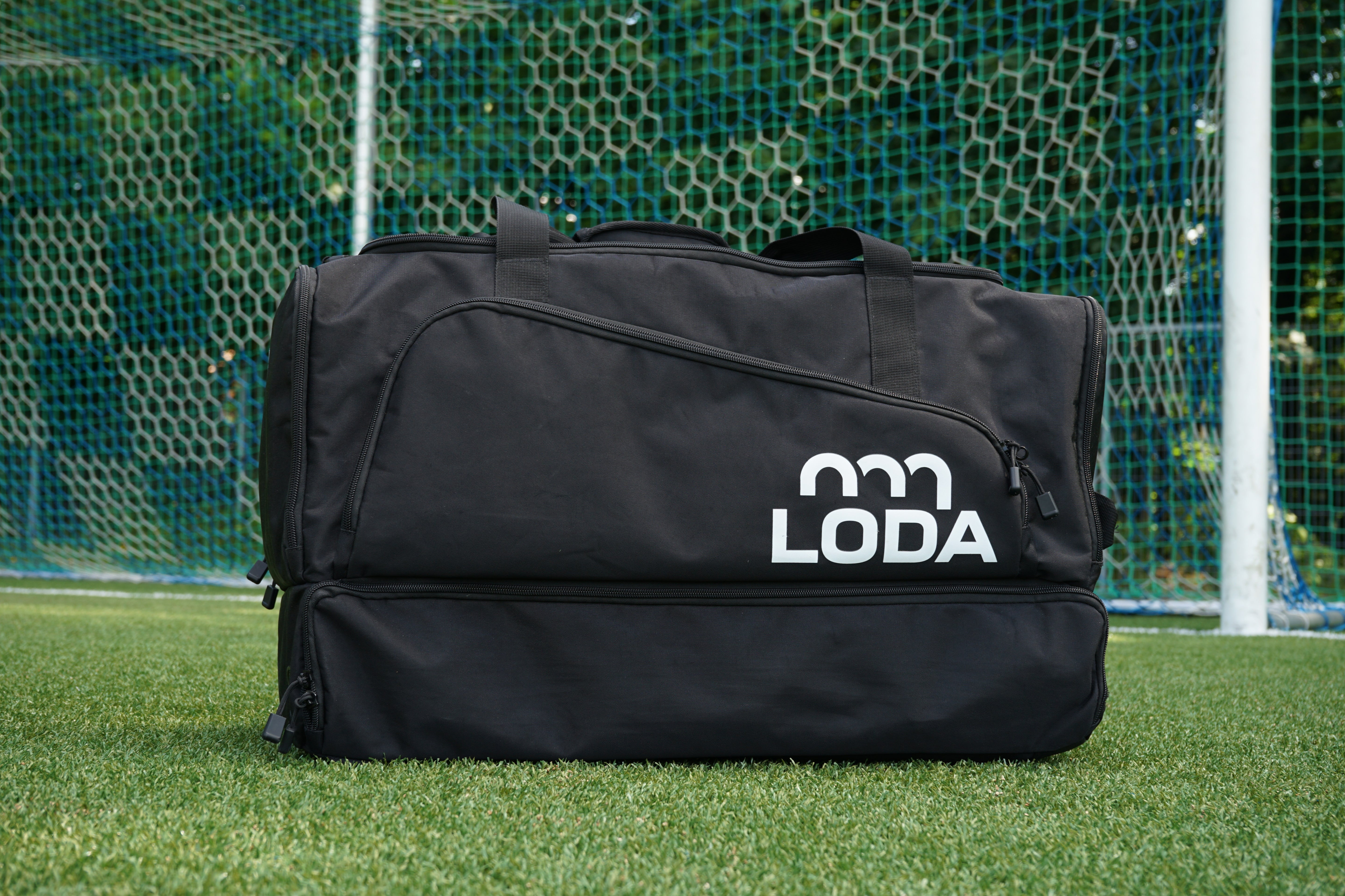 Ein großer schwarzer Seesack mit LODA-Logo liegt auf dem Rasen vor einem Fußballtornetz.