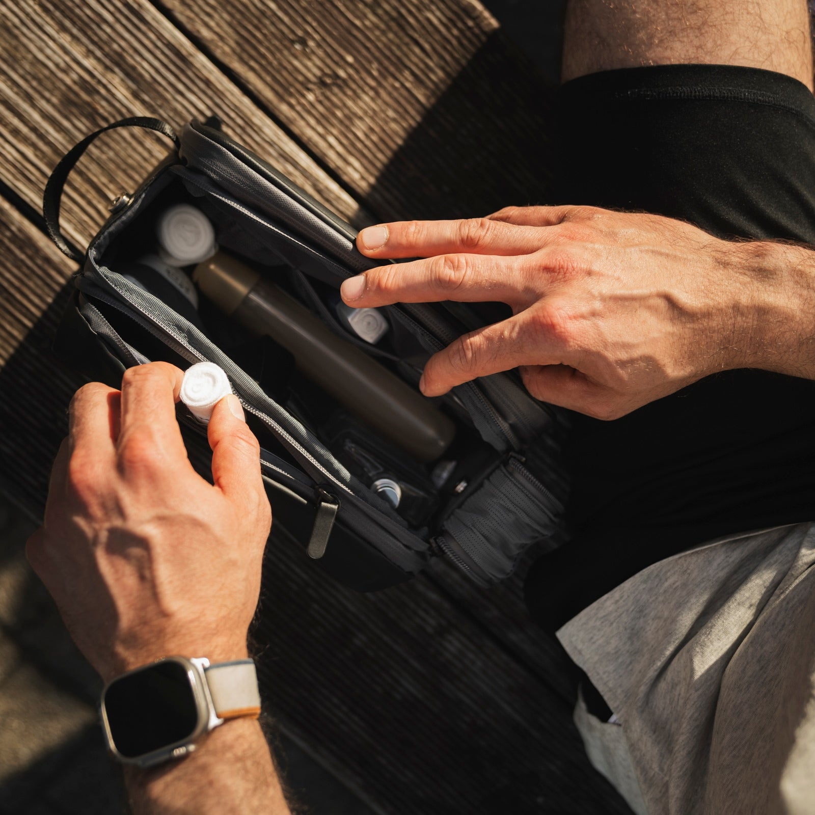Eine Person mit einer Smartwatch öffnet einen Kulturbeutel mit Flaschen, während sie auf einer Holzfläche sitzt.