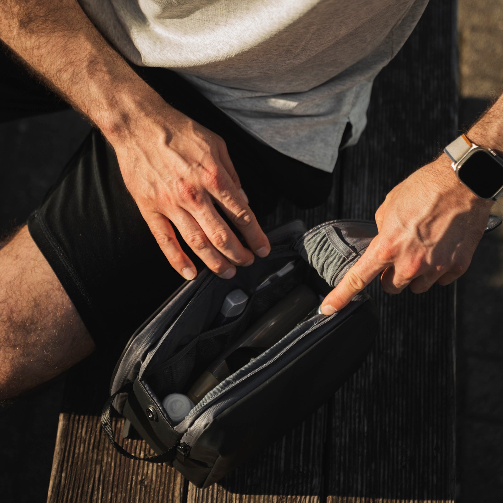 Mann in Shorts und mit intelligenter Uhr öffnet einen Kulturbeutel auf einer Holzbank im Freien.