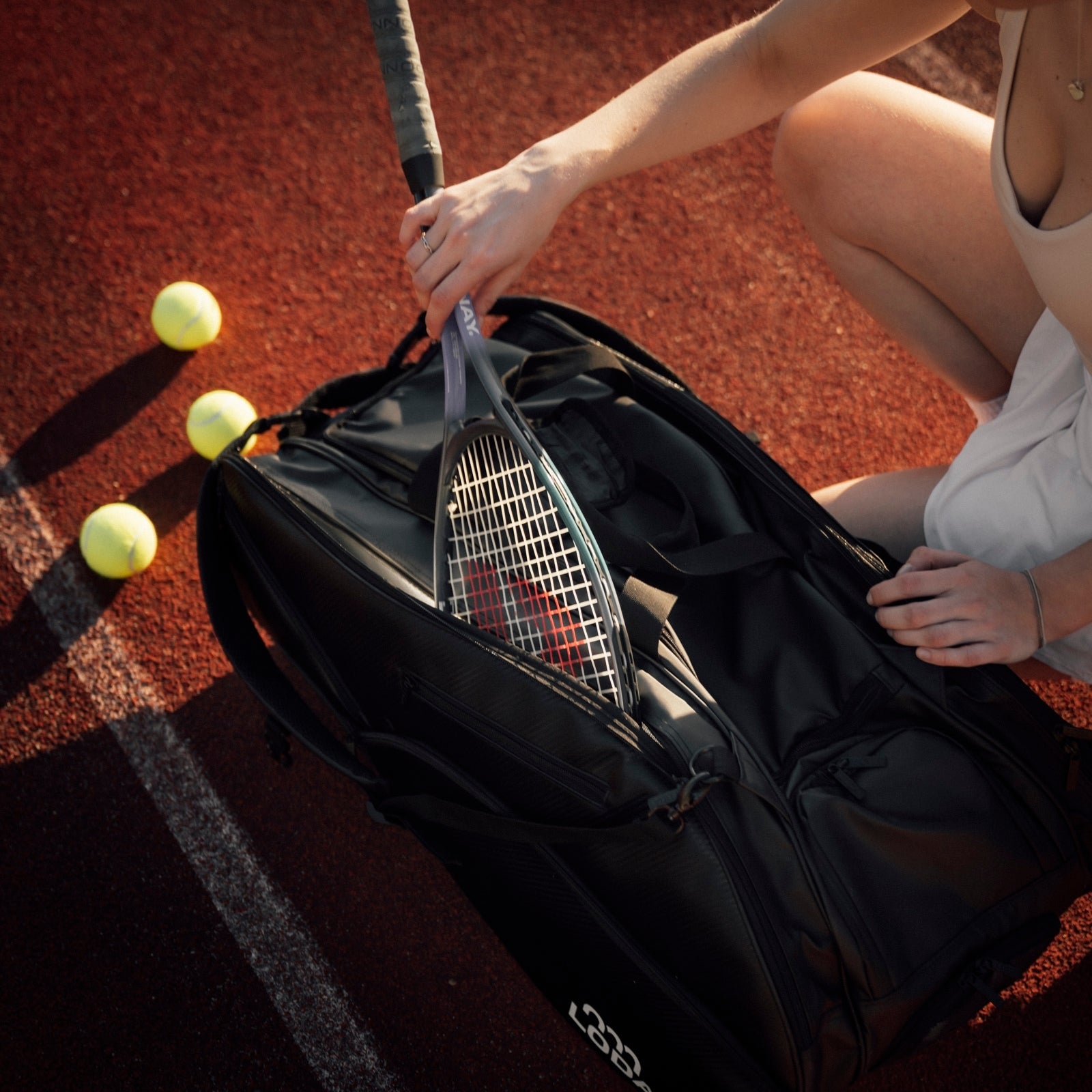 Eine Person kniet auf dem Tennisplatz und packt einen LODA Racket Pro von LODA sports in eine Tennistasche im Rucksackstil, mit Tennisbällen in der Nähe.