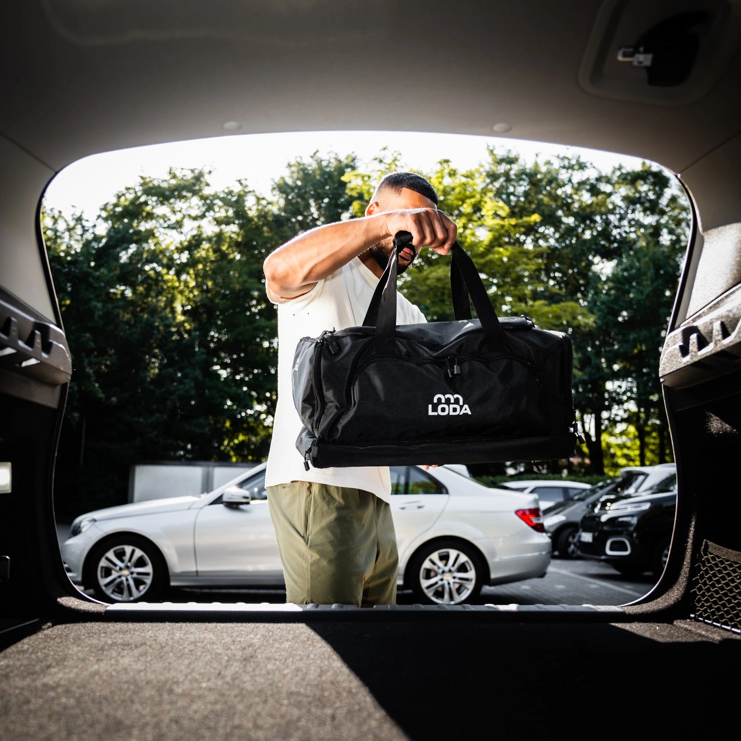 Mann, der einen schwarzen LODA-Seesack in einen Kofferraum lädt, mit geparkten Autos und Bäumen im Hintergrund.