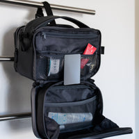 Schwarze Kulturtasche, die offen hängt und in Netztaschen organisierte Toilettenartikel vor einer weißen Wand zeigt.