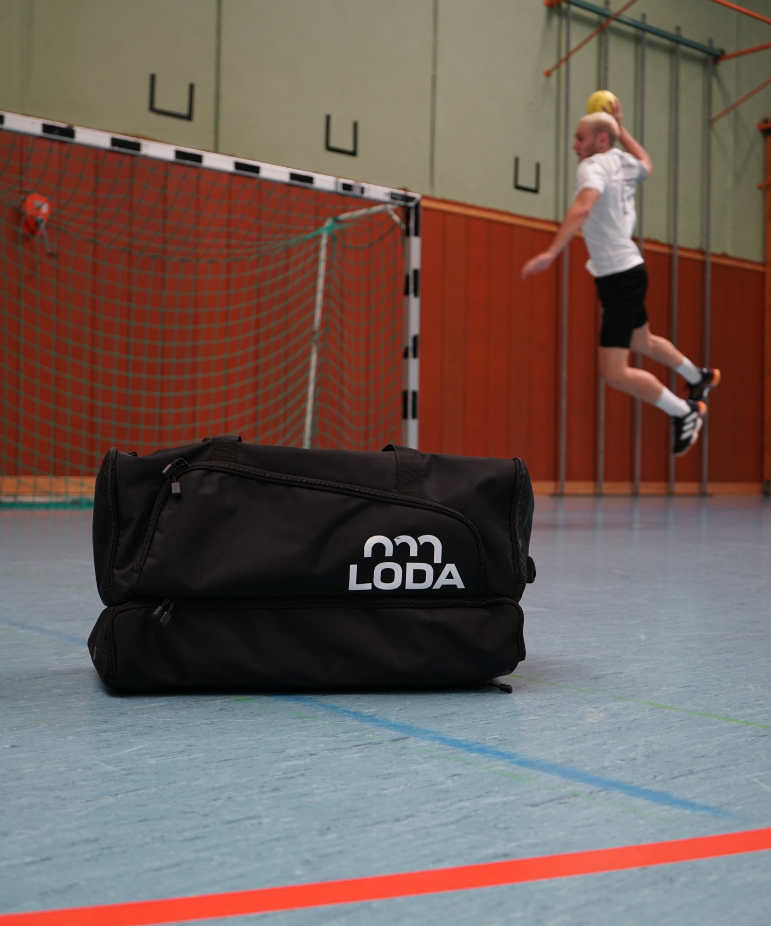 Schwarze LODA-Sporttasche im Fokus, mit einem Mann, der springt, um einen Ball auf ein Hallenhandballtor im Hintergrund zu werfen.