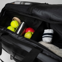 Offene Sporttasche mit Tennisbällen, Äpfeln, einer Metallflasche, einer Dose und einer gestreiften Socke darin.