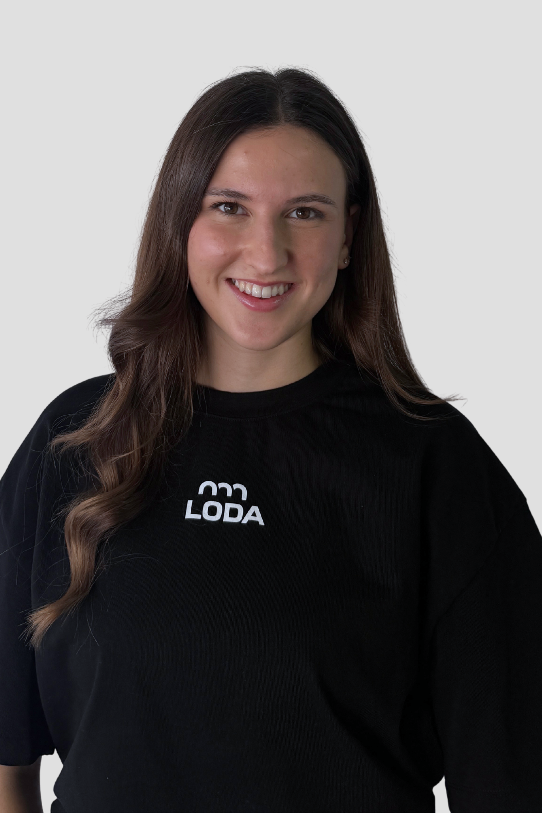 Lächelnde Frau mit langen braunen Haaren, die ein schwarzes Hemd mit der Aufschrift LODA trägt und vor einem hellen Hintergrund steht.