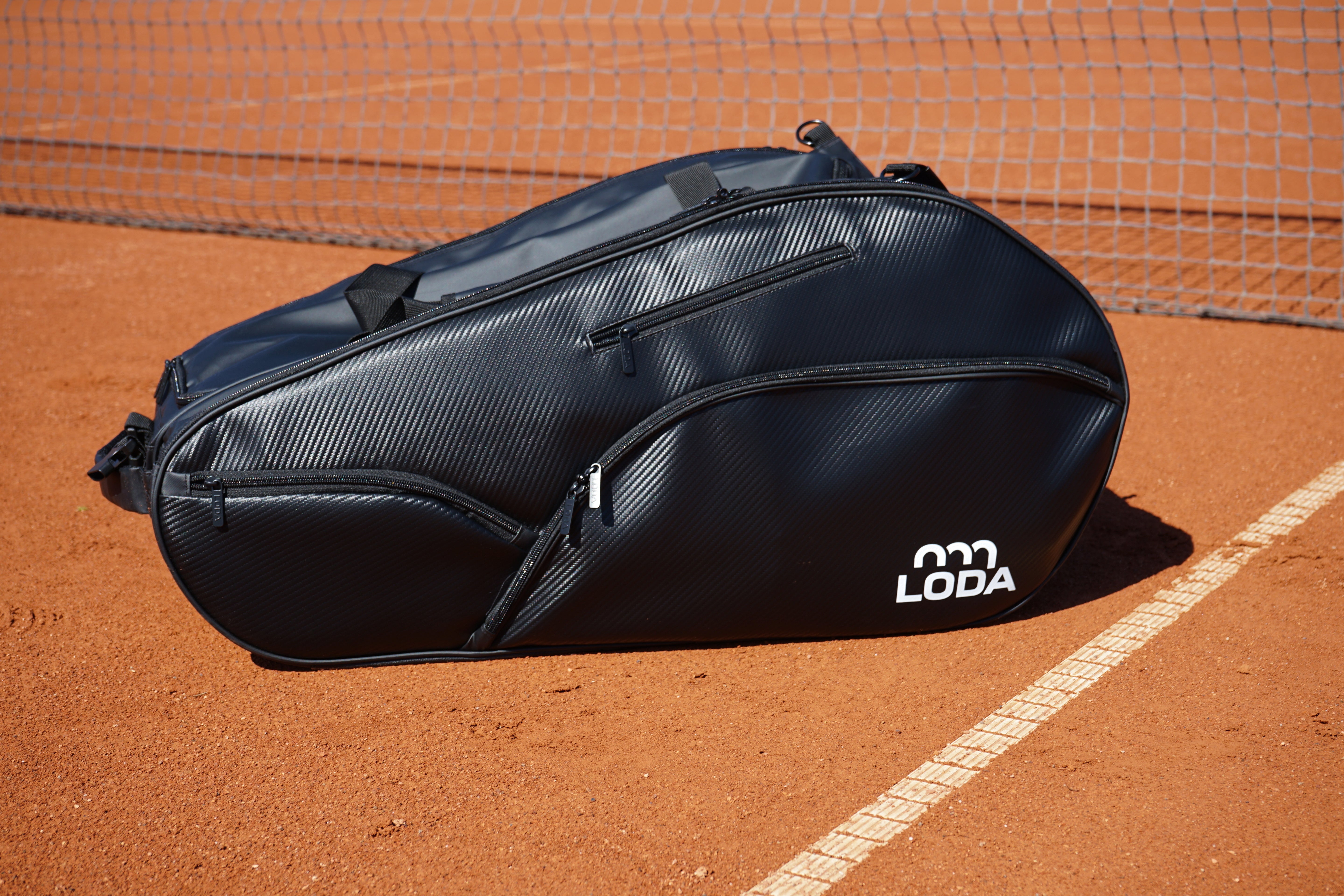 Schwarze Tennistasche mit LODA-Logo, die auf einem Sand-Tennisplatz in der Nähe des Netzes und der Begrenzungslinie liegt.