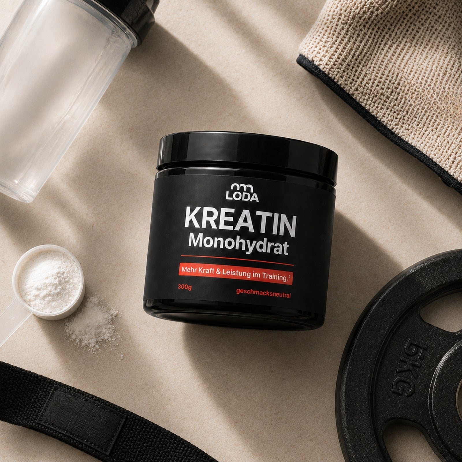 Eine Dose LODA sports LODA Kreatin Monohydrat mit einer Schaufel, einem Handtuch, einer Schüttelflasche und einer Hantelscheibe auf einer beigen Fläche.