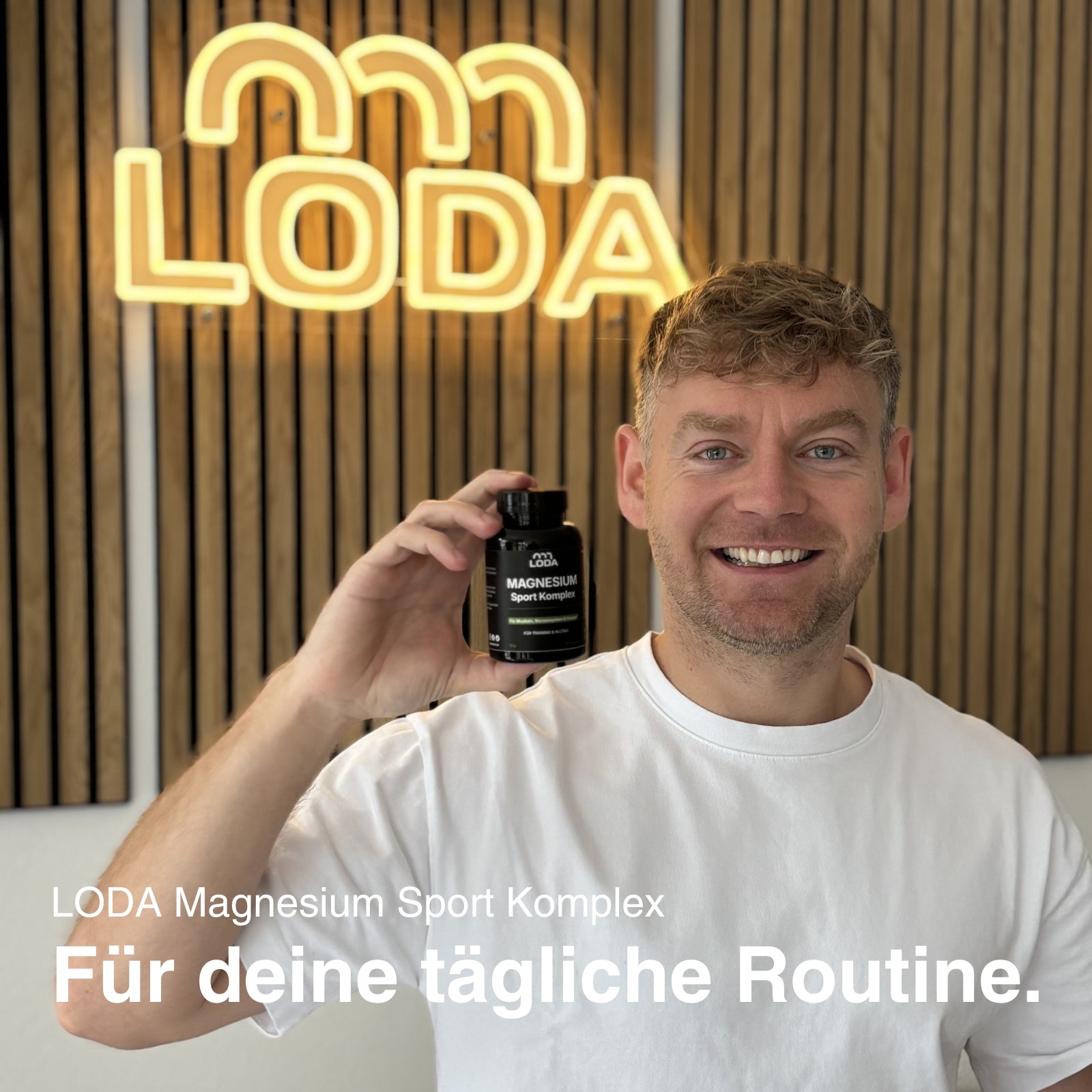 Lächelnder Mann in weißem Hemd hält eine Flasche LODA Magnesium Sport Komplex; LODA-Sportlogo und Holzpaneelwand im Hintergrund.