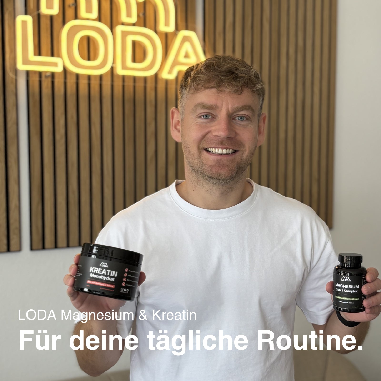 Ein lächelnder Mann im weißen Hemd hält in einem modernen Raum LODA-Sportnahrungsergänzungsmittel LODA Kreatin + Magnesium.