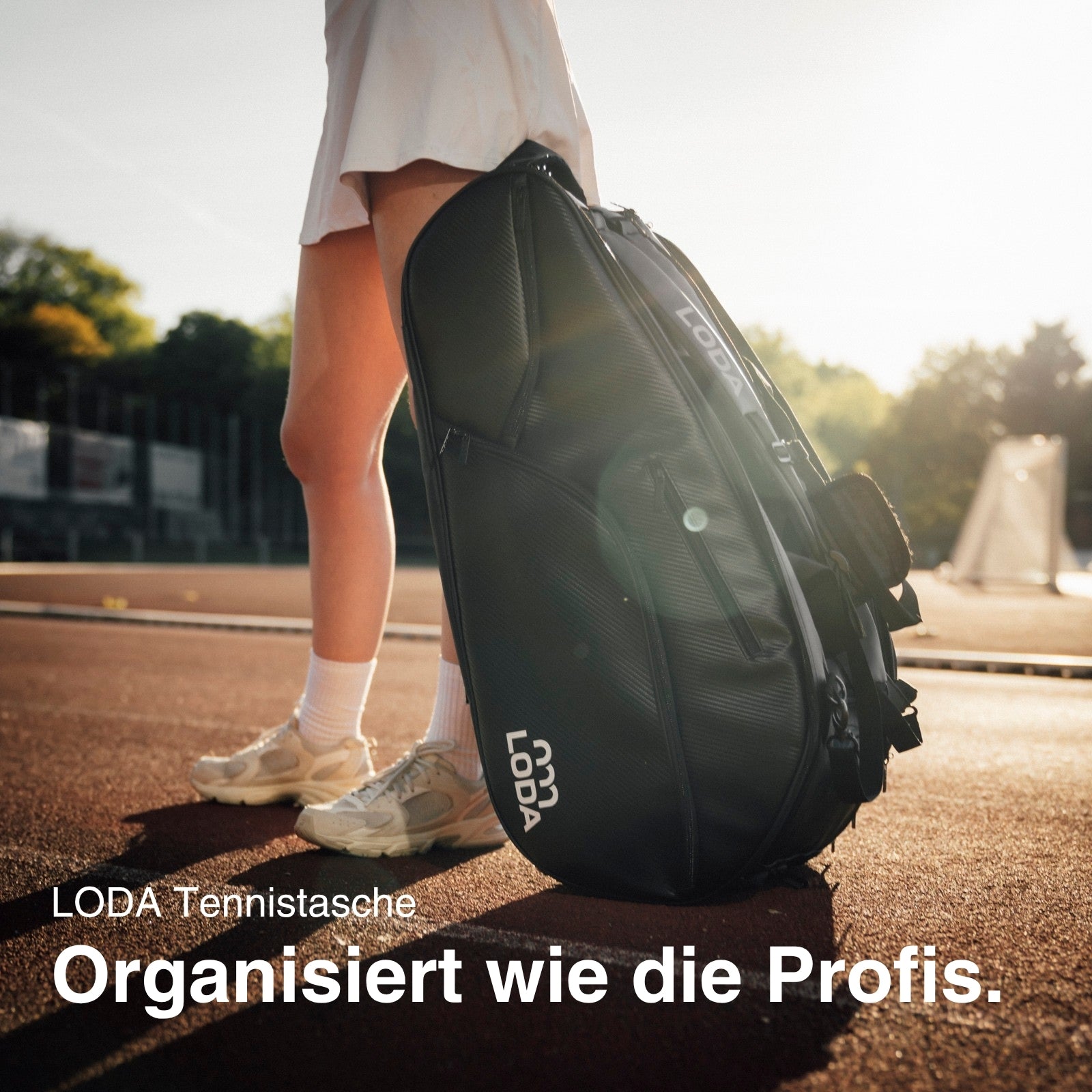 Person auf dem Tennisplatz mit einer großen schwarzen LODA sports LODA Racket Pro Tennistasche mit Rucksackfunktion, Sonnenlicht im Hintergrund, unten deutscher Text.
